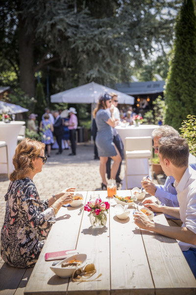 Menschen bei einem Sommerfest im Freien, drei Leute essen am Tisch im Hintergrund sind weitere Menschen und Stehtische zu sehen